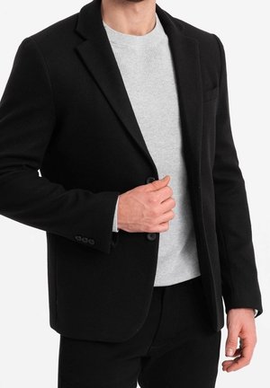 Mand iført en sort blazer over en lysegrå ribstrikket sweater, justerer blazeren med den ene hånd.