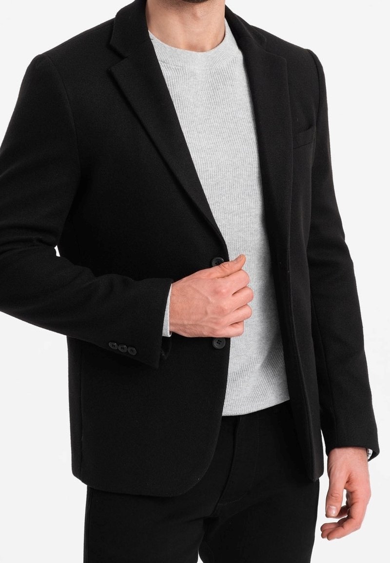 Homme portant un blazer noir par-dessus un pull côtelé gris clair, ajustant le blazer d’une main.