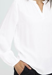 Frau in einer weißen, langärmeligen V-Ausschnittbluse mit elastischen Bündchen, teilweise mit der Hand in der Tasche einer schwarzen Hose, vor hellem Hintergrund.