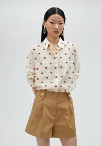 Collared button-up shirt in cream with brown polka dots, featuring two front pockets. Paired with high-waisted brown shorts.