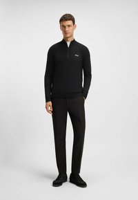 Schwarzer Reißverschluss-Pullover mit strukturierter Oberfläche, hochgeschlossenem Kragen und Logodetail, kombiniert mit schwarzen Hosen und Sneakers.