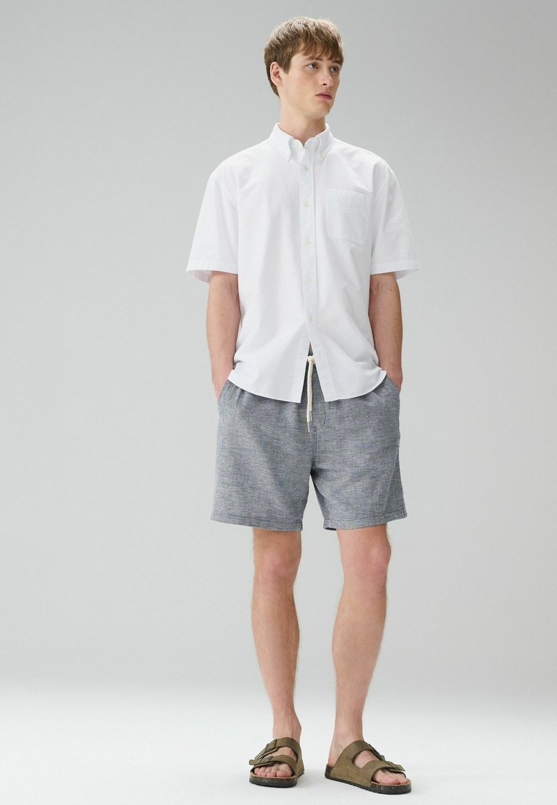 Jeune homme debout, les mains dans les poches, portant une chemise blanche à manches courtes boutonnée, un short gris et des sandales marron sur un fond uni.