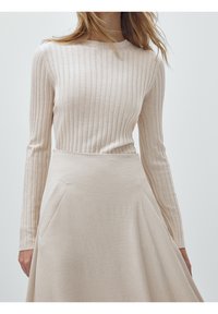 Femme portant un haut à manches longues beige côtelé rentré dans une jupe midi beige évasée taille haute avec des poches latérales.