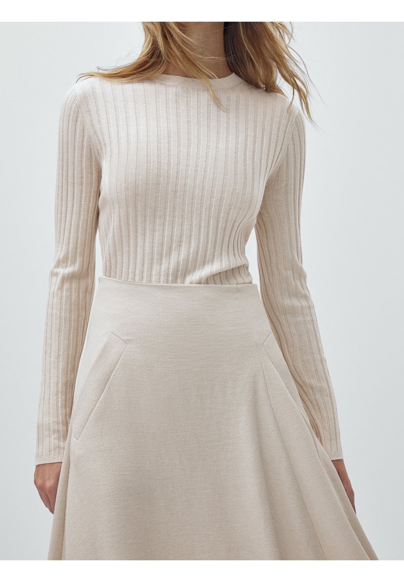 Femme portant un haut à manches longues beige côtelé rentré dans une jupe midi beige évasée taille haute avec des poches latérales.