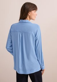 Camicia a maniche lunghe di colore azzurro chiaro con colletto, dotata di una piega sul retro e polsini abbottonati. Tessuto morbido e testurizzato, design semplice.