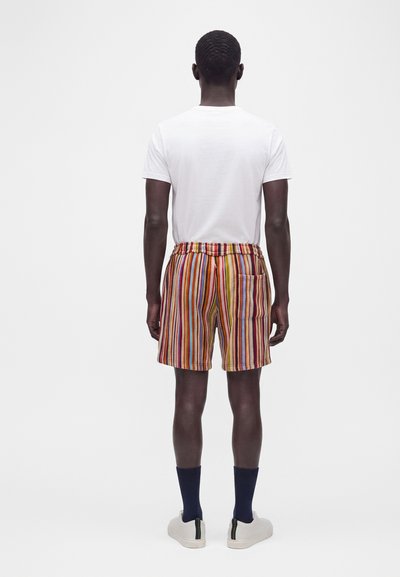 Homme debout, dos tourné, portant un t-shirt blanc, un short à rayures verticales colorées avec une poche arrière, des chaussettes noires et des baskets blanches sur fond uni.