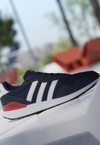 Zapatilla Adidas azul marino con rayas blancas y detalle rojo en el talón, colocada sobre una superficie blanca al aire libre con fondo borroso.