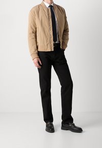 En tanfärgad bomberjacka med ribbade muddar, dragkedja och sidofickor, kombinerad med svarta jeans och svarta loafers, vilket skapar en avslappnad look.