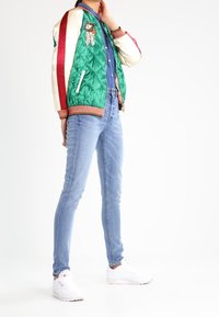 Chaqueta bomber verde y crema con rayas rojas, detalles bordados, combinada con jeans ajustados azul claro y zapatillas blancas.