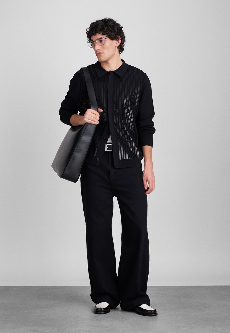 Schwarze Strickjacke mit Streifenmuster, schwarze Hose und schwarze Schultertasche. Glänzende Akzente auf der Strickjacke und kontrastierende Schuhe.