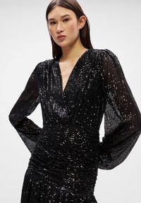 Vestido preto com lantejoulas, apresentando mangas longas e bufantes, decote em V profundo e detalhes franzidos, criando um efeito texturizado e cintilante.