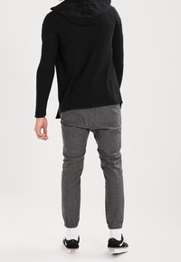 Uomo che indossa un hoodie nero, pantaloni grigi con risvolto, calze bianche e sneaker neri senza lacci, in piedi su uno sfondo bianco semplice.