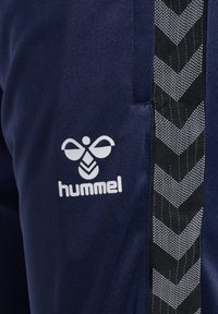 Granatowe sportowe ubrania z białym logo „hummel” oraz czarnym bocznym panelem z wzorem jodełki w kolorze białym. Teksturowane wykończenie tkaniny.