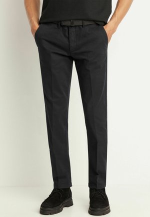 Hombre con pantalones negros de corte ajustado con cinturón negro tejido, camisa negra y zapatos de ante negros con cordones, manos en los bolsillos.