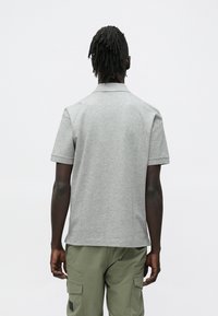 Mężczyzna o ciemnej skórze i dredach, zwrócony tyłem, ubrany w jasnoszare polo z krótkim rękawem i oliwkowe spodnie cargo na białym tle.