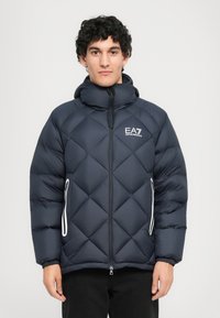 Férfi, fekete steppelt pufidzsekiben, kapucnival, elülső cipzárral, fehér cipzáras oldalsó zsebekkel és EA7 Emporio Armani logóval a mellkasán.