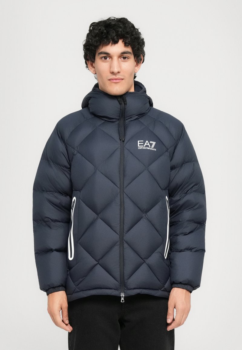 Férfi, fekete steppelt pufidzsekiben, kapucnival, elülső cipzárral, fehér cipzáras oldalsó zsebekkel és EA7 Emporio Armani logóval a mellkasán.