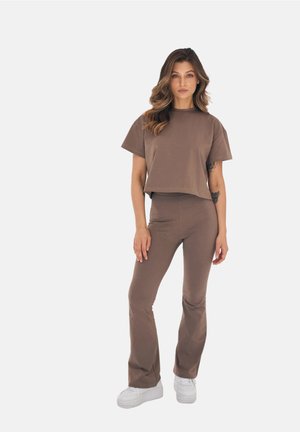 Kobieta stojąca, ubrana w brązowy krótki top z krótkim rękawem i dopasowane, rozkloszowane spodnie oraz białe platformowe trampki, jasne tło.