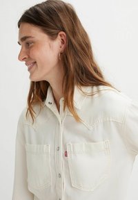 Camisa de botón color crema con dos bolsillos en el pecho, costuras en contraste y cierres a presión plateados. Tela suave con un yugo de estilo western.