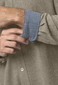 Hemd mit grau-braunem Körper und blauen Chambray-Manschetten, ausgestattet mit einer Knopfleiste und sichtbarer Stoffstruktur.