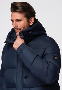 Granatowa kurtka puchowa z wysokim kołnierzem, wykonana z lekkiego materiału, z widocznymi szwami, zatrzaskami i naszywką z logo na rękawie.