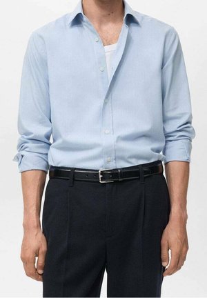 Homme portant une chemise bleu clair boutonnée avec manches retroussées, un tee-shirt blanc, une ceinture noire et un pantalon noir sur un fond uni.