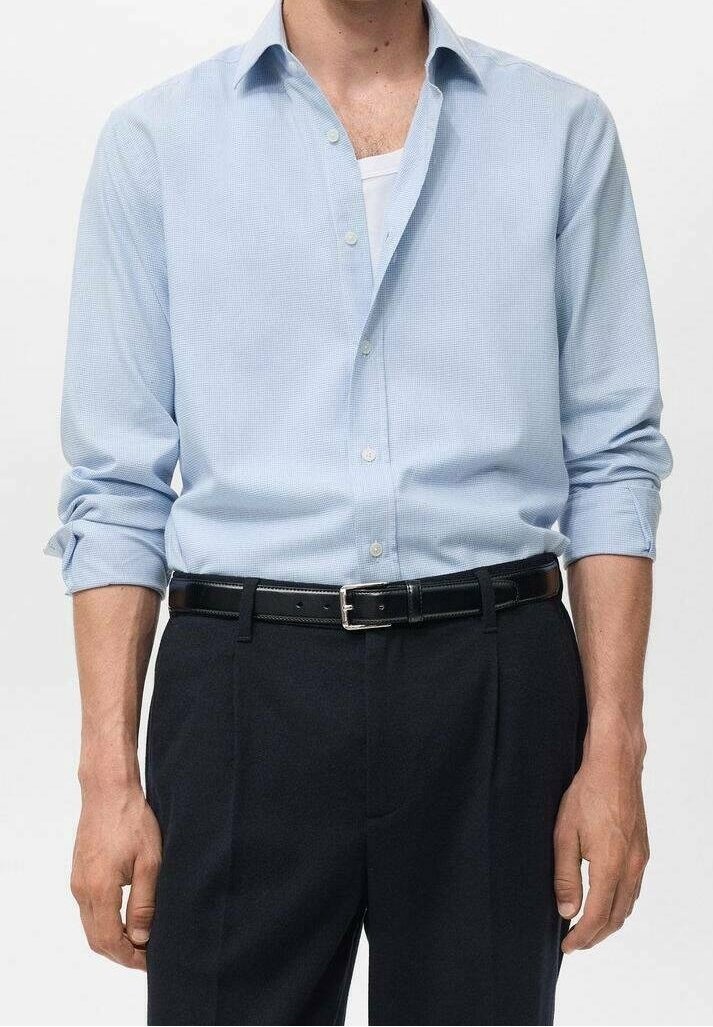 Homme portant une chemise bleu clair boutonnée avec manches retroussées, un tee-shirt blanc, une ceinture noire et un pantalon noir sur un fond uni.