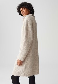Maglione lungo beige a maglia con texture a coste, colletto alto e spacchi laterali, indossato sopra leggings neri. Il modello è in piedi di profilo.