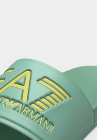 Ανοιχτό πράσινο σανδάλι slip-on με ανάγλυφο κίτρινο λογότυπο «EA7 Emporio Armani» στο λουρί, εμφανισμένο σε απλό λευκό φόντο.