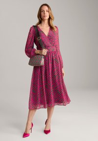 Rosa Langarmkleid mit spiralförmigem Muster, tailliertem Bund und fließendem Rock. Hält eine gemusterte Handtasche, kombiniert mit rosa High Heels.