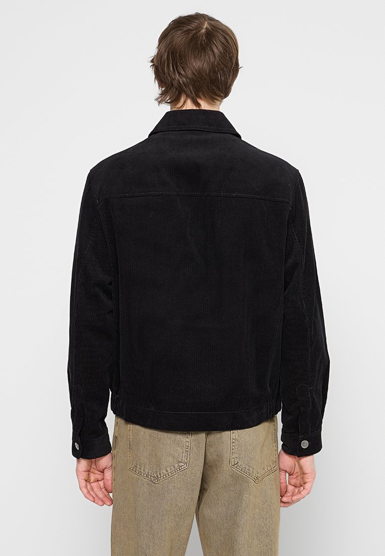 Veste en velours côtelé noire avec un col classique, des manches longues et des poignets boutonnés ; présente une coupe droite et un détail de tige au dos.