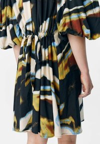 Robe avec un motif abstrait vibrant en noir, or, bleu et beige. Taille élastique avec un détail à nouer et des manches bouffantes.
