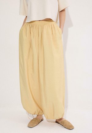 Lõdvad, kõrge vöökohaga kahvatukollased püksid, millel on elastne vöökoht ja nööriga alumised servad, kantud heledate pruunide kootud libisemiskindlate kingade ja lühema beežika ülaosaga.