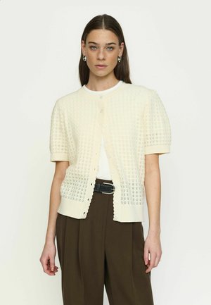 Ženska s ravnimi rjavimi lasmi nosi kremasto kratko pleteno jopico s kratkimi rokavi čez belo majico in ohlapne rjave hlače s črnim pasom, nevtralno ozadje.