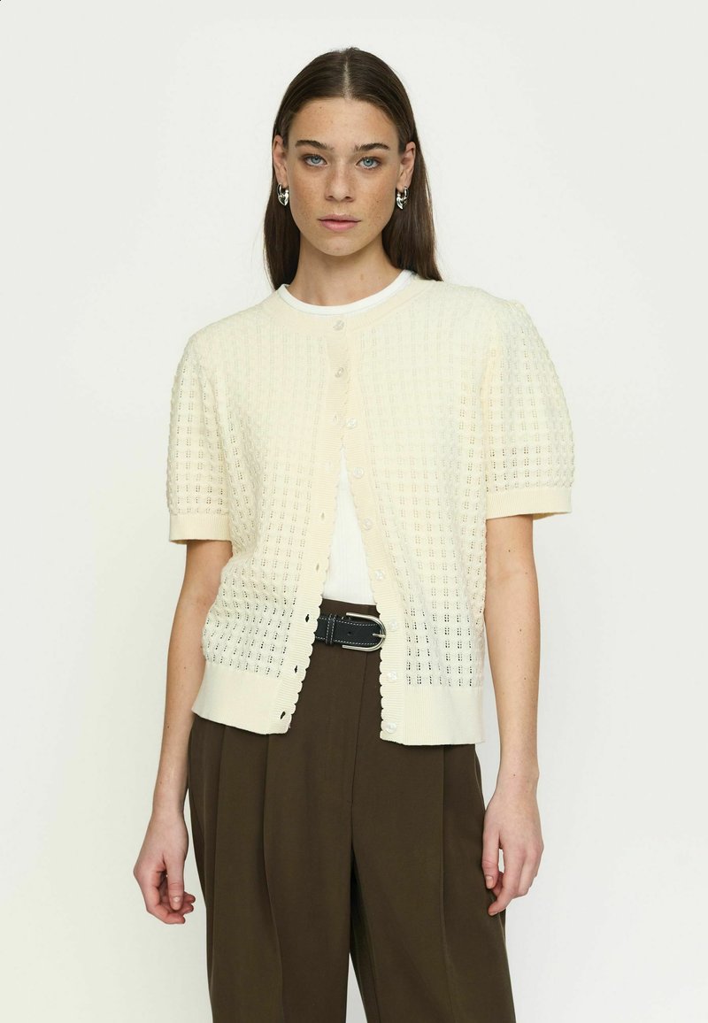 Moteris su tiesiais rudais plaukais dėvi kreminės spalvos trumpomis rankovėmis megztą kardiganą ant baltos palaidinės ir laisvus rudus kelnes su juodu diržu, neutraliame fone.