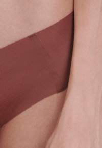 Bordeaux seamless slip met een gladde textuur. Elastische tailleband, lage snit en een nette afwerking aan de randen. Huidskleurige hand in het frame.