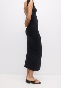 Robe noire moulante sans manches avec un dos décolleté ; longueur mi-mollet avec une texture lisse. Associée à des sandales noires à imprimé léopard.