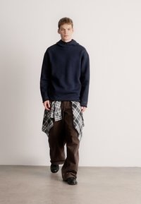 Tmavě modrá mikina s kapucí, hnědé široké kalhoty a šedá károvaná košile uvázaná kolem pasu. Model má na sobě černé boty a stojí na neutrálním pozadí.