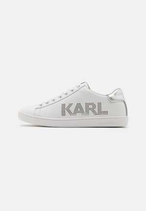 Zapatilla blanca de cuero con detalles perforados que forman "KARL". Presenta acentos plateados en el talón y los ojales de los cordones. Suela plana de goma.
