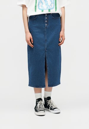 Osoba nosi modrú rifľovú sukňu strednej dĺžky so zapínaním na gombíky a predným rozparkom, doplnenú bielymi ponožkami a čiernymi vysokými teniskami.