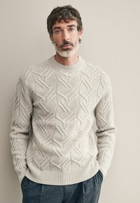 Hellgrauer Strickpullover mit strukturiertem Zopfmuster, dicker Rundhalsausschnitt und geripptem Saum. Kombiniert mit dunkelgrauen, maßgeschneiderten Hosen.