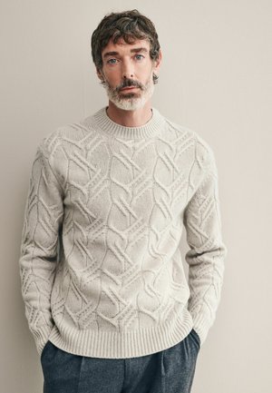 Hellgrauer Strickpullover mit strukturiertem Zopfmuster, dicker Rundhalsausschnitt und geripptem Saum. Kombiniert mit dunkelgrauen, maßgeschneiderten Hosen.