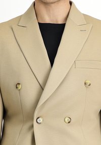 Blazer doppiopetto beige in tessuto texture, con rever a laccetto e tre bottoni decorativi di colori alternati. Tasche a scomparsa sul petto.