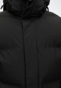 Gros plan sur une doudoune noire avec un col haut et des coutures apparentes, montrant le nom de la marque discrètement embossé sur la poitrine.
