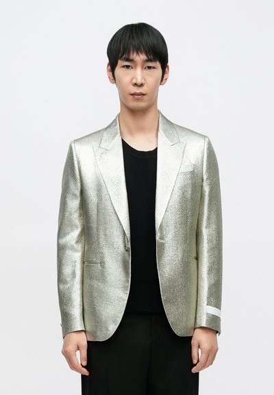 Blazer argenté métallique avec revers à crans, fermeture à un bouton et poches. Porté par-dessus un t-shirt noir à col rond, associé à un pantalon noir.