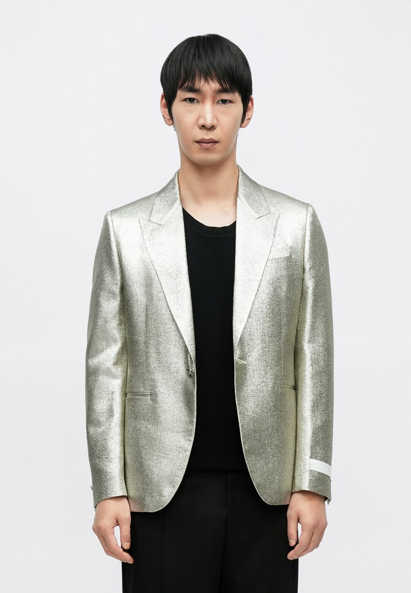 Metaliczna srebrna marynarka z wyciętymi klapami, pojedynczym zapięciem na guzik i kieszeniami. Nosi się ją na czarnej koszulce z okrągłym dekoltem, w połączeniu z czarnymi spodniami.