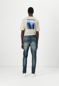 Camiseta beige con un estampado gráfico en la parte posterior, combinada con jeans azul oscuro y zapatillas blancas. El conjunto tiene un corte relajado y un diseño casual.