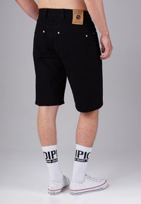 Black shorts with a slim fit, featuring a leather label on the waistband and copper-tone hardware. Worn with white socks and sneakers.