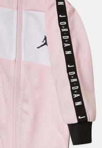 Rosa-weiße Jackenärmel mit schwarzem Bund, schwarzer vertikaler Streifen am Arm mit dem Text "Jordan" und einem schwarzen Jumpman-Logo auf einem weißen Feld.