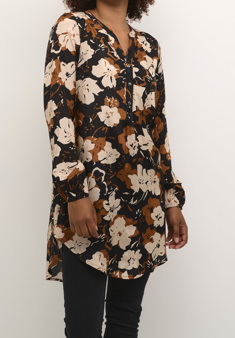 Femme portant une tunique noire à manches longues avec un grand imprimé floral beige et marron, col boutonné, associée à un pantalon noir.